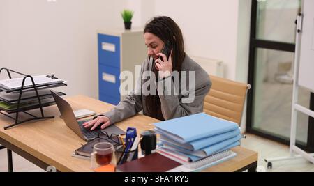 Focused young woman in gray blazer talking on smartphone while working on laptop at office desk with folders, paperwork, and stationery in bright Stock Photo