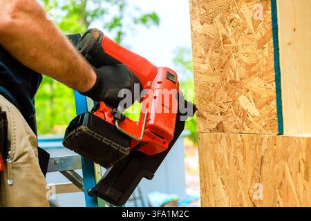 Carpenter stands on ladder using air nail gun to secure wood panels together at construction location Stock Photo