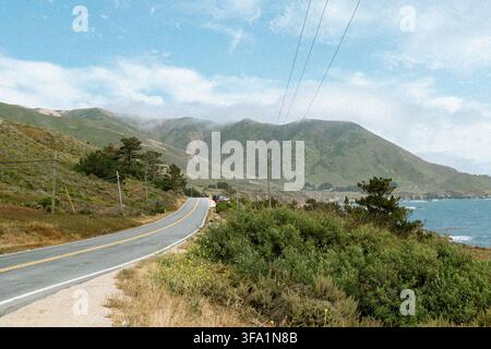 A winding coastal highway curves along green mountains and the ocean, with fog settling over the peaks above Stock Photo
