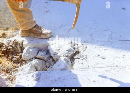 Worker uses pickaxe to break concrete at construction site dust rises from ground. Stock Photo