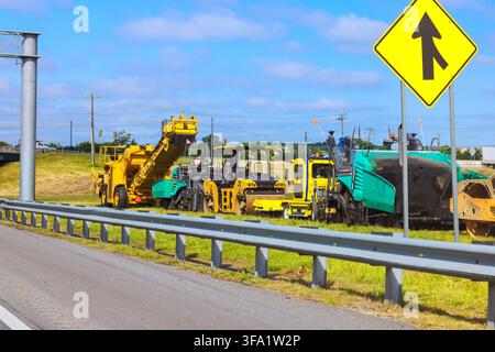 Several large construction vehicles are parked on grass beside highway wait for work on site. Stock Photo