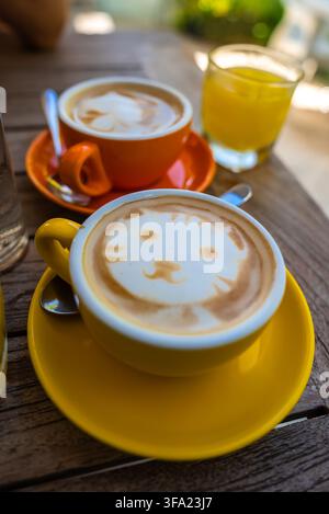 Yellow ceramic cup of cappuccino featuring creative cat latte art rests on a wooden table beside an orange coffee cup and orange juice. Stock Photo