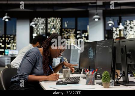 Programmer taking notes at office desk, reviewing artificial intelligence system metrics. Tech company employee writing down info, monitoring performance of machine learning APIs Stock Photo