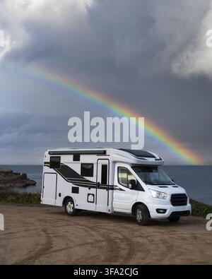 View of a white camper van parked on a dirt road near the sea at Capo Mannu with a vibrant rainbow in the stormy sky in Sassari, Sardinia, Italy. Stock Photo