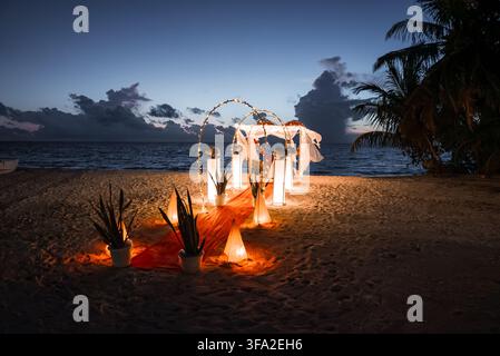 An illuminated wedding arch with an aisle and dinner table is set up on a sandy beach on Dhigurah island in Maldives during twilight. Stock Photo