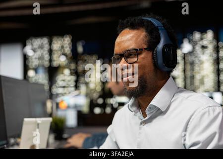 Smiling technician listening music in startup office, optimizing code for performance and scalability, doing quality assurance. Happy IT worker wearing audio headphones using computer Stock Photo