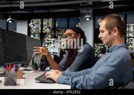 Cheerful tech startup intern asks questions to senior programmer coding advanced AI algorithms and machine learning models enhancing automation. Smiling worker requests help from manager in office Stock Photo