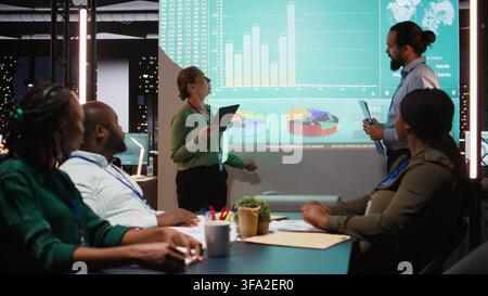 Diverse staff from an enterprise sharing data analytics and insights for a new project agenda during late night corporate meeting. Workers discuss about resources allocation. Camera A. Stock Photo