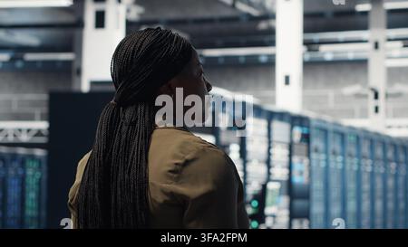 Woman walking through data center server rows, checking equipment for system stability. IT professional overseeing server farm, monitoring infrastructure mainframes, camera B Stock Photo
