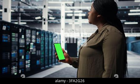 Software developer using app on mockup phone to check server hub energy consumption. African american woman using green screen smartphone in data center to do gear checkup, camera A Stock Photo