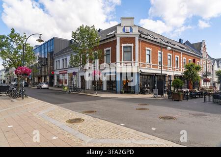 View of a street corner featuring a historic red brick building, a modern glass facade, and vibrant flower baskets under a bright blue sky in Kristian Stock Photo
