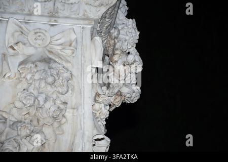 Close-up crop of a carved marble ribbon bow and floral wreath relief flanking a larger sculptural group, isolated against deep black shadow. Stock Photo