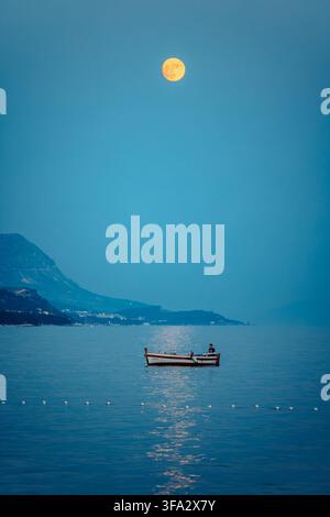 View of a small boat with a fisherman floating on the calm Adriatic Sea under a bright full moon with distant mountains in the background in Brela, Sp Stock Photo