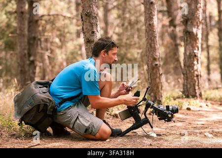 Outdoor photography session in forest with a man using a camera and smartphone for creative work during sunny day Stock Photo
