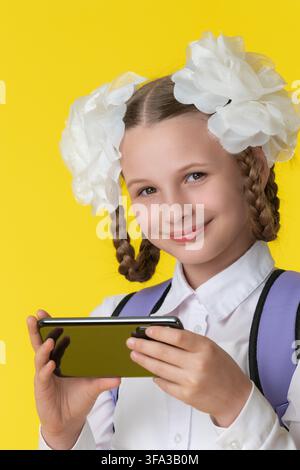 Happy student with braided hair, white bows, and backpack holds smartphone. Girl in uniform smiles at camera on yellow background. Concept for modern Stock Photo