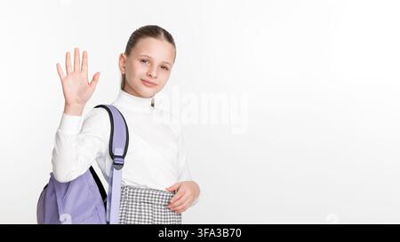 Happy school girl with purple backpack waves hand in greeting. Student portrait with friendly expression on white studio background. Ample copy space Stock Photo