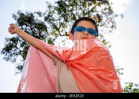 Child dressed as a superhero with a plastic cape and blue sunglasses, looking up with one arm raised, representing imagination, strength, and dreams o Stock Photo