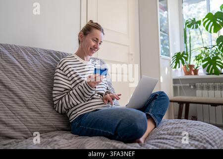 Smiling relaxed woman sitting on sofa at home using laptop and credit card for online shopping. Stock Photo