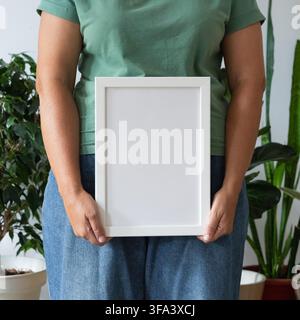Woman in green t-shirt and blue jeans holding empty frame in space filled with plants against white wall. Modern minimalist design Stock Photo