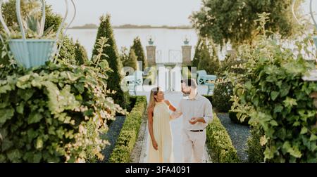 A young couple enjoys a romantic walk in a beautifully landscaped garden. The sun sets, casting a warm glow over the serene surroundings. Stock Photo