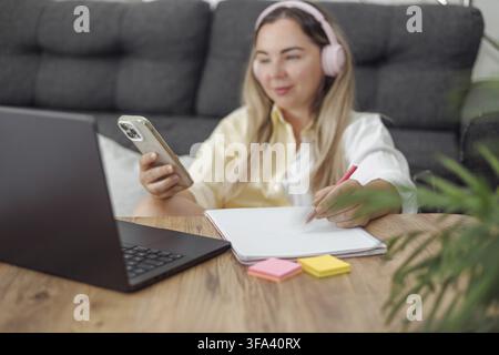 Young blond European woman with headphones using smartphone and laptop at wooden table with notebook and sticky notes indoors Stock Photo
