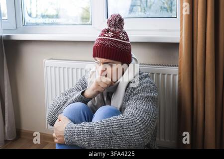 Freezing young man in winter clothes coughs and warms up near the radiator at home Stock Photo