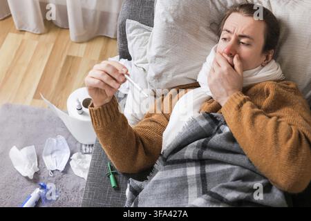 Sick man with flu checking thermometer while lying on couch at home surrounded by wipes and medication Stock Photo