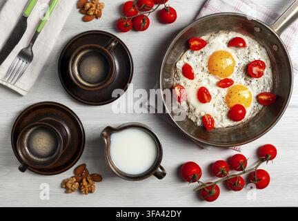 Two fried eggs with cherry tomatoes in a pan, two cups of coffee, milk, nuts and cherry tomatoes branches are composing a delicious breakfast on a whi Stock Photo