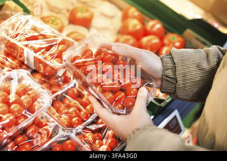 Close-up of a person's hands holding a clear plastic container of fresh cherry tomatoes in a grocery store Stock Photo