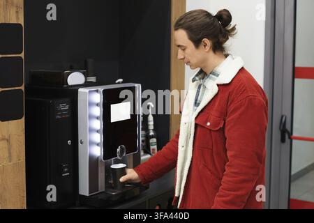 Young man wearing a red jacket uses a coffee machine in an office or waiting area for a hot drink Stock Photo