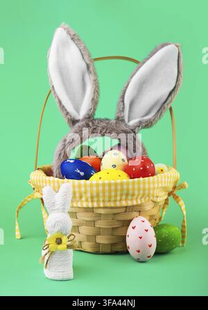 Festive Easter basket filled with colorful painted eggs and fluffy bunny ears. A white string bunny and decorated eggs sit nearby against a bright gre Stock Photo