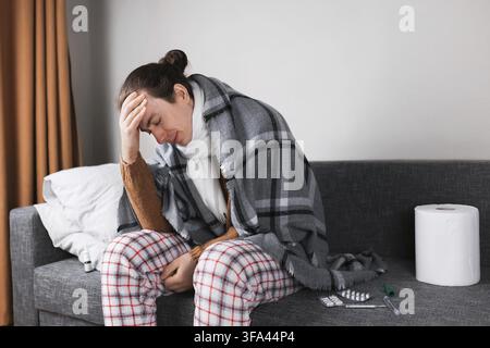 Sick man in pajamas and blanket suffering from headache and flu while sitting on sofa at home Stock Photo