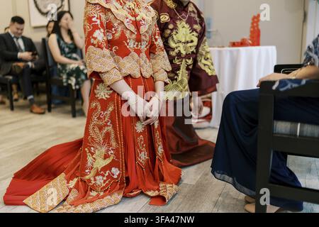 Bride and groom dressed in ornate red and gold traditional Chinese attire kneel during a tea ceremony, with family and guests present in a formal indo Stock Photo