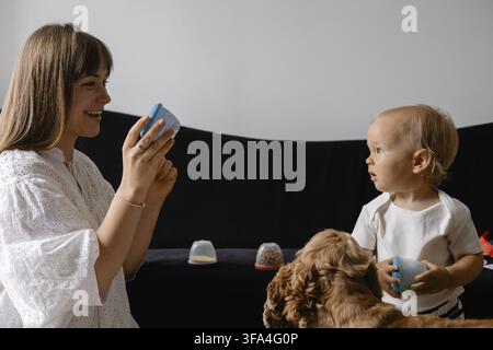 A mother and baby interact with colorful cups while a dog observes, highlighting playtime and companionship Stock Photo