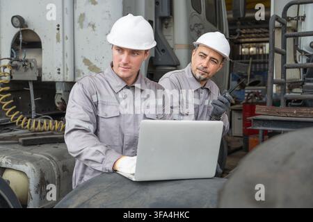 Two male mechanics reviewing equipment maintenance plans on a laptop in an industrial workshop. Ideal for automotive repair, management, and teamwork Stock Photo