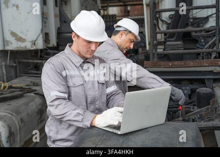 Two technicians working on machinery with a laptop at a site for efficient maintenance. Ideal for concepts in engineering, technology, and industrial Stock Photo