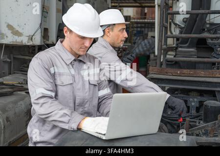 Two technicians working on machinery with a laptop at a site for efficient maintenance. Ideal for concepts in engineering, technology, and industrial Stock Photo