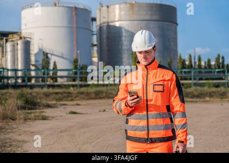 Construction worker in safety gear using smartphone at industrial site for communication. Ideal for themes of industry, technology, safety, and workfo Stock Photo