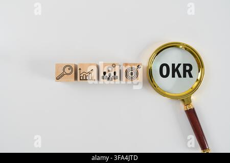 Magnifying glass focusing on OKR beside wooden blocks with key, chart, team and target icons, illustrating objectives, key results, strategy, performa Stock Photo
