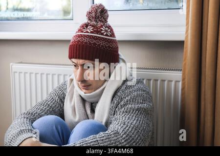Freezing young man wearing a winter hat and scarf is sitting near the radiator at home and waiting for heat Stock Photo