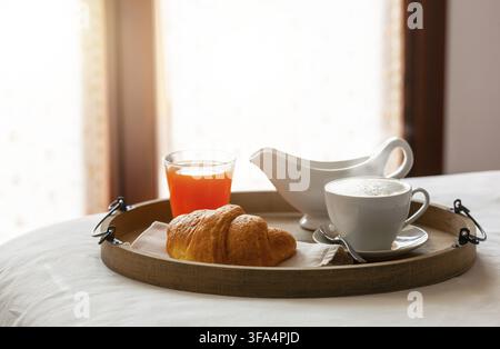 Rich breakfast with croissant, cappuccino, orange juice and milk jug is served on a wooden tray on a bed in a hotel room with natural light entering f Stock Photo