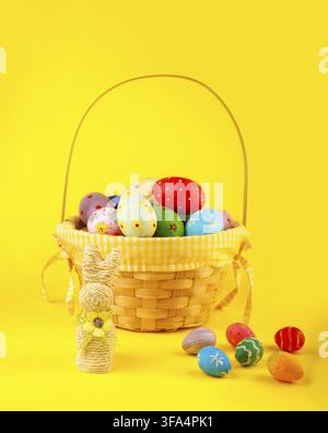 A festive vertical shot of a wicker basket overflowing with colorful hand-painted Easter eggs and a straw bunny figurine, set against a bright, solid Stock Photo
