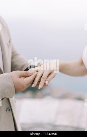 A man in a beige suit gently places an engagement ring on a womans finger, both hands visible, with a soft blurred outdoor background suggesting a rom Stock Photo