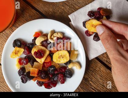 Hand picking crispy dried banana chips from a colorful mix of nuts and dried fruits, beautifully arranged on a white plate atop a wooden table, accomp Stock Photo