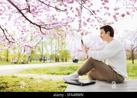 A satisfied freelancer takes a break outside from working from home. The young man sits with his laptop under blooming cherry trees and smiles in his Stock Photo
