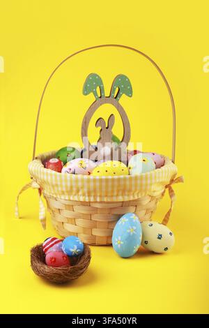 Colorful hand-painted Easter eggs in a wicker basket with a wooden bunny ornament and a small nest against a vibrant yellow background. Cheerful sprin Stock Photo
