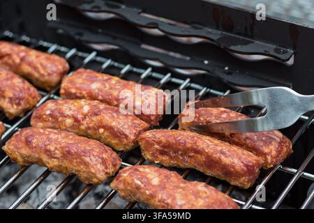 Traditional Romanian mici skinless minced meat rolls grilling on a hot barbecue grate with a metal fork turning one over. Stock Photo