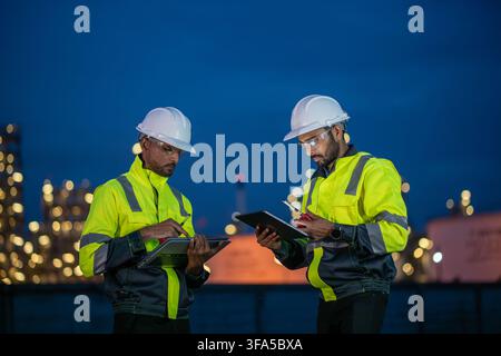 Engineers wearing safety gear, including hard hats examining survey tablet standing industrial facility gas task or oil refinery. Stock Photo