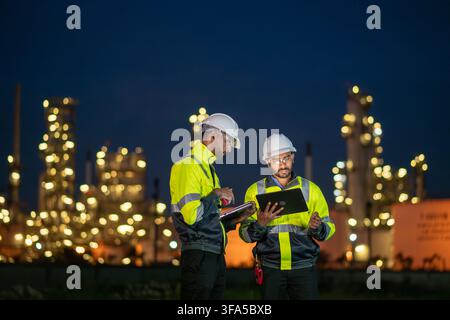 Engineers wearing safety gear, including hard hats examining survey tablet standing industrial facility gas task or oil refinery. Stock Photo