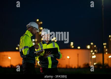 Engineers wearing safety gear, including hard hats examining survey tablet standing industrial facility gas task or oil refinery. Stock Photo